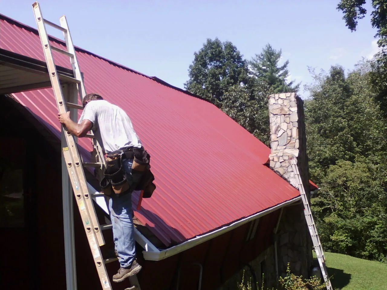 Roofer on a ladder beside a red metal roof and stone chimney — Soffit & Fascia Repair by Roofing Brothers in Beaver Crossing